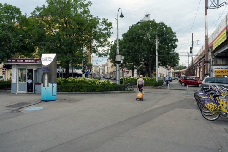 Person geht mit einem Einkaufswagen über einen Platz in Wien-Landstraße, Bäume, Laternen, Bahntrasse, Fahrräder, Trinkbrunnen, Imbissstand