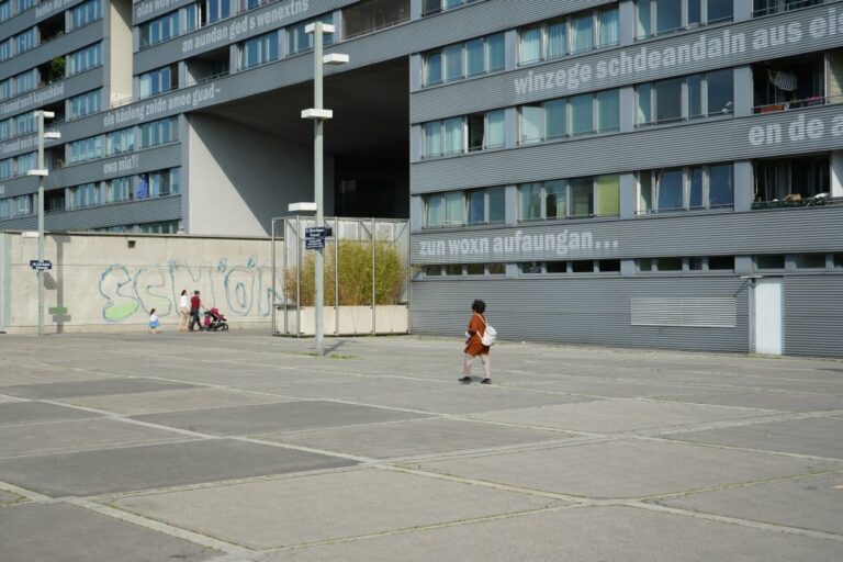 Frau und Familie mit Kinderwagen vor einem breiten Wohnhaus in Wien-Donaustadt