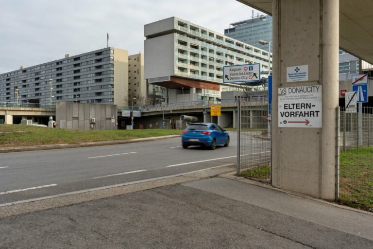 Auto fährt auf einer Straße vor einem modernen Stadtviertel in Wien, Schilder rechts: "VS Donaucity", "Elternvorfahrt"