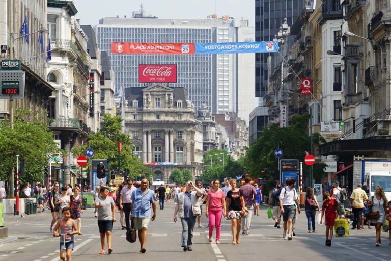 Fußgängerzone im Zentrum Brüssels, hinten Werbung für Coca Cola, dahinter ein Hochhaus, Transparent "Place au piéton! Voetganger koning!"