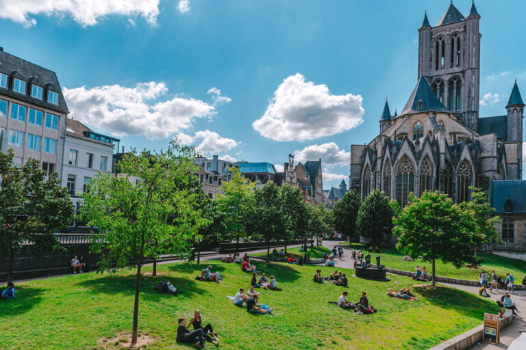 Leute liegen auf dem Rasen neben Bäumen in einem Park im Zentrum der Stadt Gent, rechts eine Kirche (Parochiekerk Sint-Niklaas)