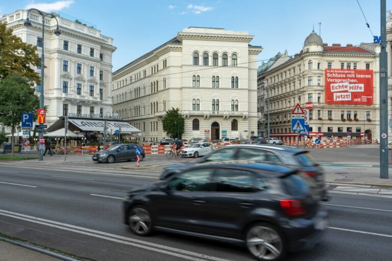 Autos fahren am Universitätsring, dahinter das Café Landtmann und die Zentrale der SPÖ mit Plakat