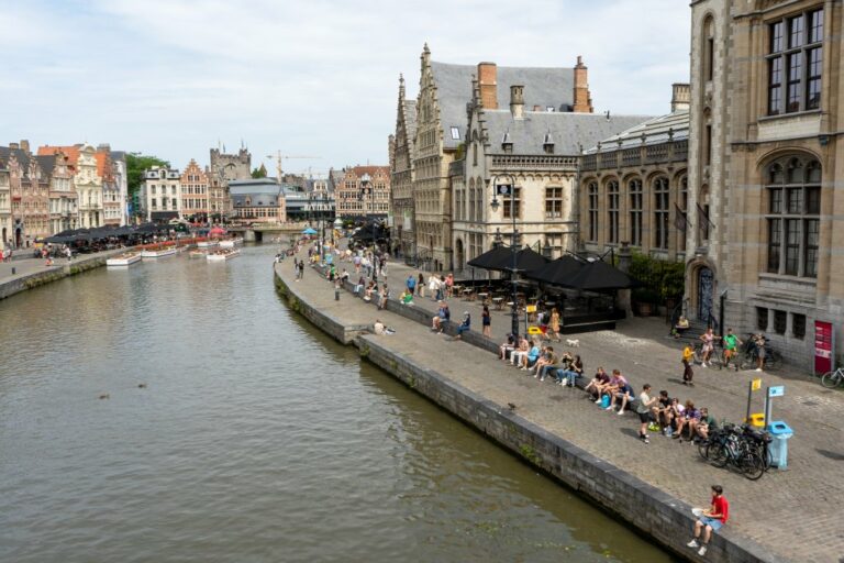 Leute sitzen am Ufer eines Flusses, dahinter alte Gebäude
