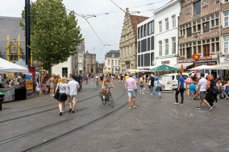 Innenstadt von Gent in Belgien, Frau fährt ein Rad, viele Leute gehen herum, Gastgärten, Geschäfte, alte Häuser, Baum, Straßenbahnschienen, Oberleitungen