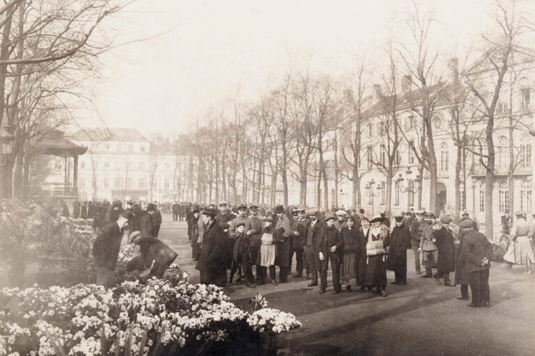 altes Foto eines Blumenmarkts, in der Mitte viele Menschen, hinten Bäume und Gebäude