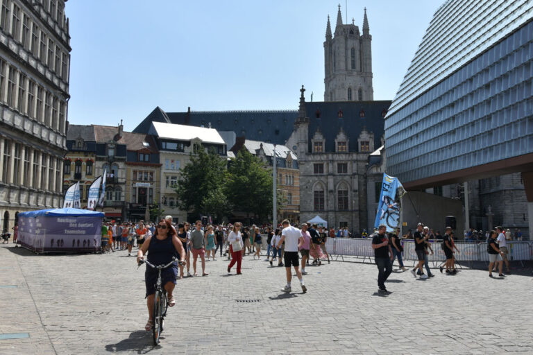 Radfahrerin und viele Leute auf einem autofreien Platz in Gent, links das Rathaus, in der Mitte die Tuchhalle und die Kathedrale, rechts eine moderne Veranstaltungshalle