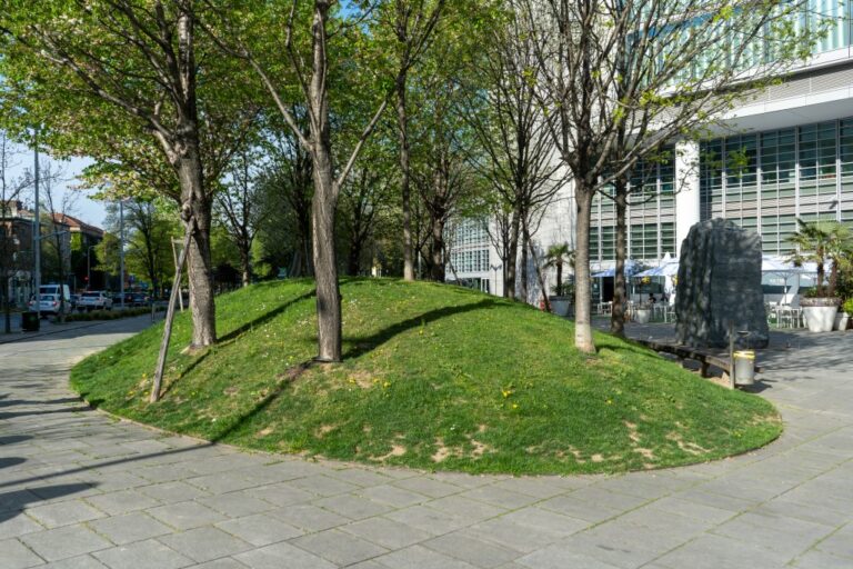 Hügel mit Gras und Bäumen, rundherum gepflasterte Fläche, rechts ein modernes Bürohaus