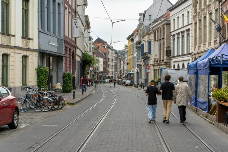 drei Männer gehen auf der Fahrbahn einer Straße im Zentrum von Gent, rechts ein Gastgarten, links parkendes Auto und abgestellte Räder