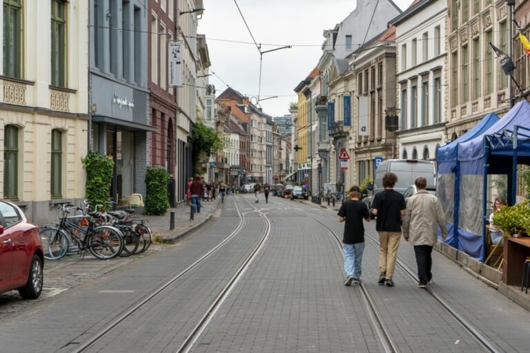 drei Männer gehen auf einer Straße im Zentrum von Gent, rechts ein Gastgarten, links parkendes Auto und abgestellte Räder