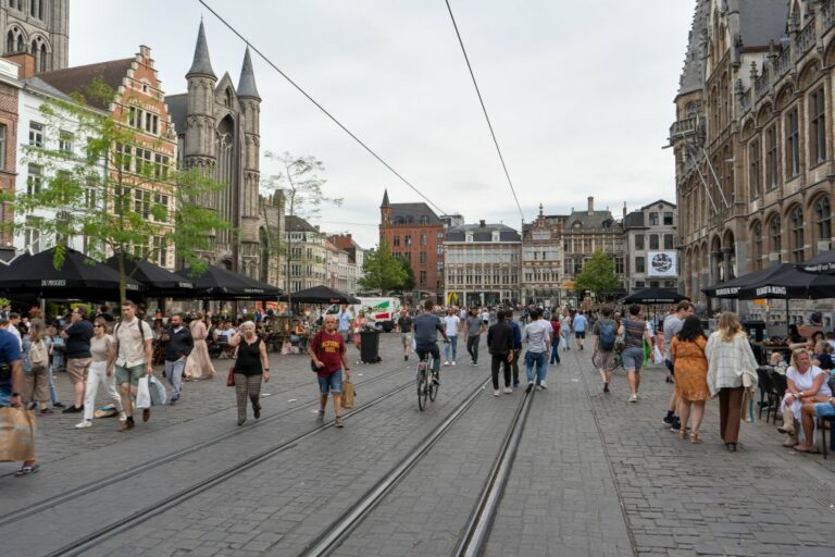 Fußgängerzone mit vielen Menschen umgeben von historischen Gebäuden, Türme einer Kirche, Straßenbahnschienen, Oberleitungen. Bäume, Gastgärten