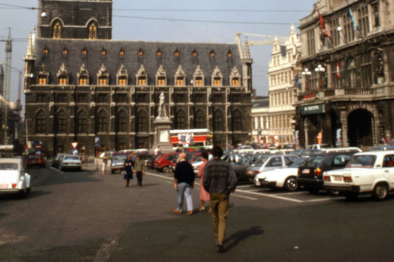 Personen gehen über einen Stadtplatz, auf dem viele Fahrzeuge abgestellt sind, Tuchhalle, Belfried, Rathaus, Theater