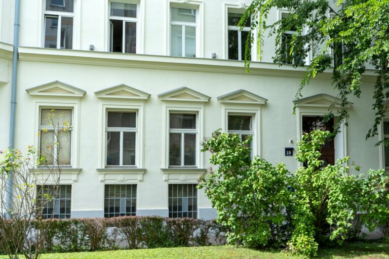Erdgeschoßfassade eines renovierten Gründerzeithauses im 4. Bezirk in Wien, davor eine Grünfläche mit Sträuchern und Baum