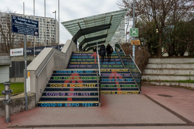 Stufen zu einer überdachten Promenade in einem modernen Stadtviertel in Wien, Hinweis zu COVID-19 Schnelltests in mehreren Sprachen