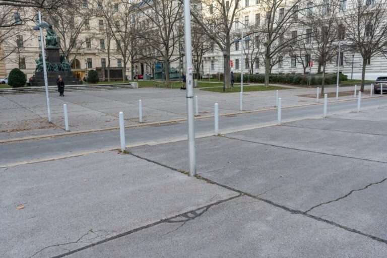 Platz in Wien, Bäume, Poller, Fahrbahn, Denkmal, Personen, Straßenlaternen