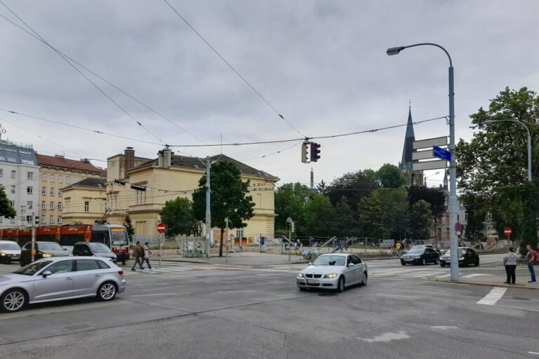 Kreuzung an der Ecke Neubaugürtel/Stollgasse in Wien, fahrende Autos, Straßenlaternen, Kirchturm, historischer Pavillon