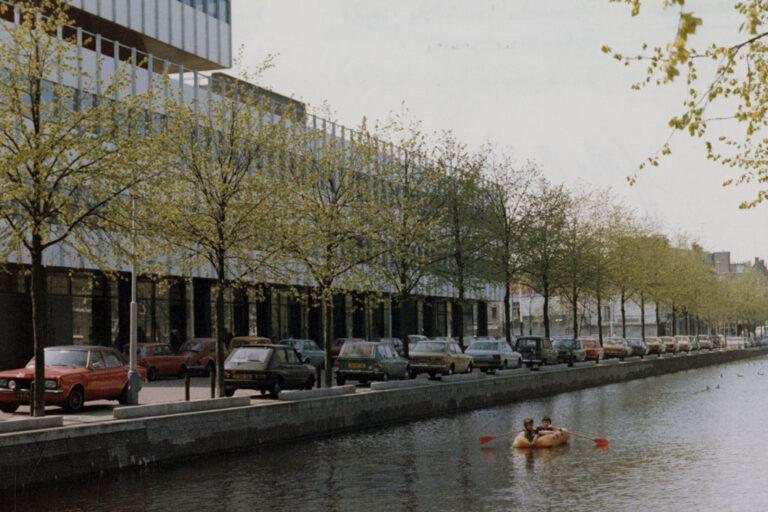 Kinder auf einem Schlauchboot in einem Kanal in Den Haag, im Hintergrund ein Bürohaus, Bäume, Autos