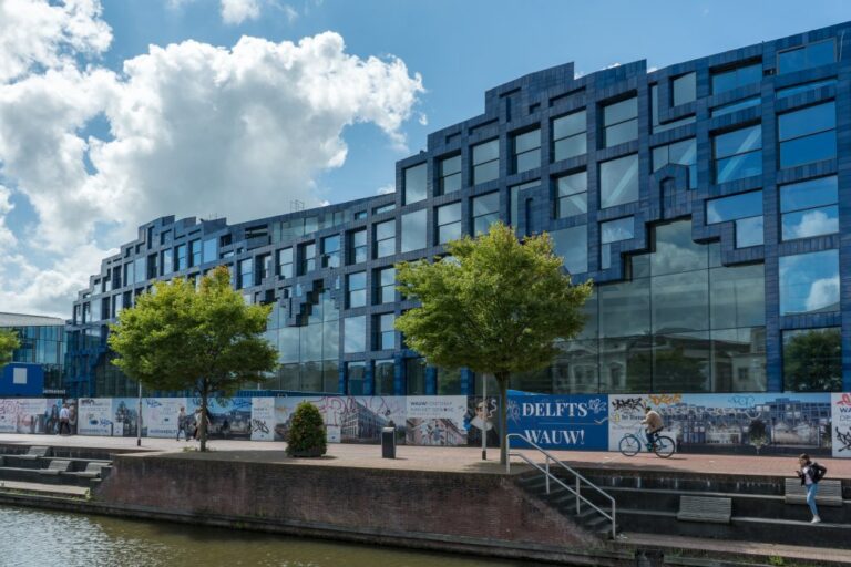 langes Gebäude mit blauer moderner Fassade, davor Bäume und eine Gracht