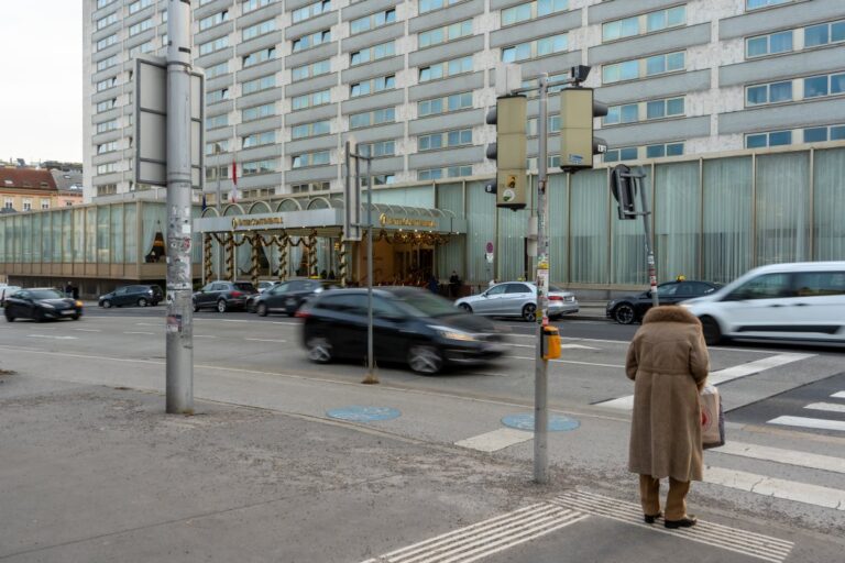 Person mit Wintermantel wartet an einer Ampel, Autos fahren vor einem Hochhaus vorbei, Radweg