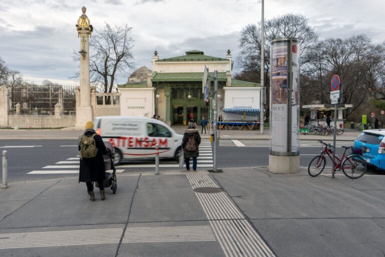 zwei Personen stehen an einer roten Ampel, Lieferauto fährt über die Straße, Stationsgebäude im Jugendstil, Blindenleitsystem auf Asphalt, Fahrrad, Verkehrsschild, Litfaßsäule, Bäume