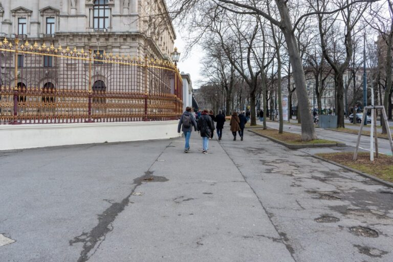 Personen gehen auf einem Gehsteig der Wiener Ringstraße, links Zaun und Neue Hofburg