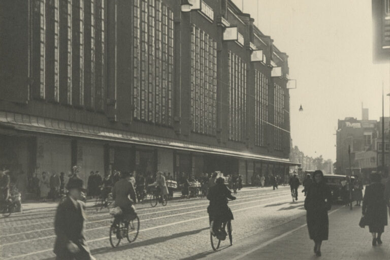 Fußgänger und Radfahrer in einer Einkaufsstraße vor einem großen Kaufhaus mit hohen Fensterfronten