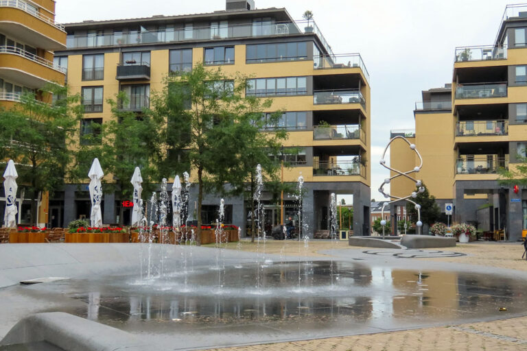 Wasserspiele, Brunnen, Platz vor Wohnhäusern, Bäume, Schirme, Kunstwerk