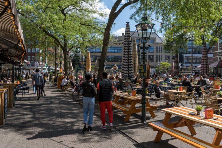 Gastgarten auf einem Platz, Leute, Laterne, Bäume, Tische, Markise, Schirme