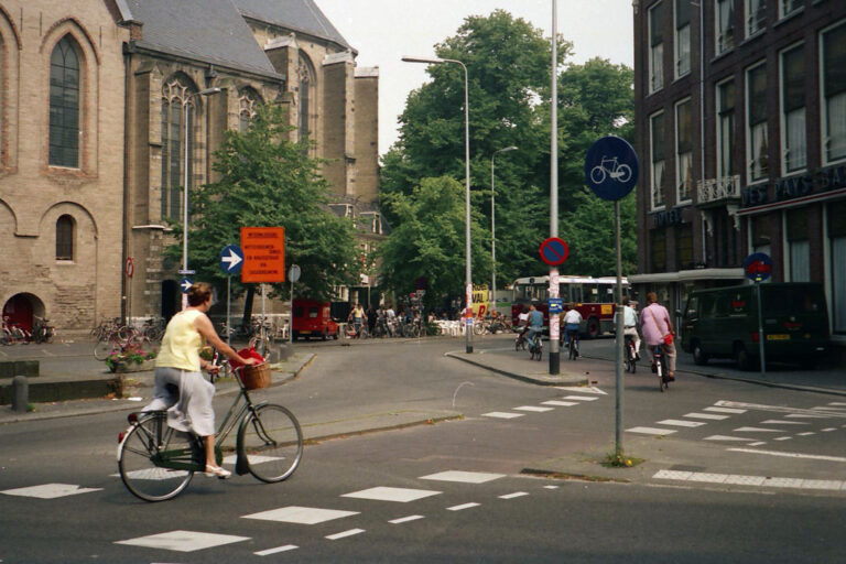 Frau fährt auf einem Fahrrad auf einem Radweg über eine Straße im Zentrum von Utrecht, im Hintergrund eine Kirche und Bäume