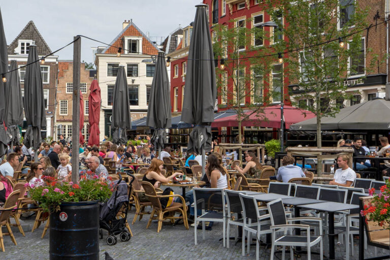 Leute sitzen in einem Gastgarten auf einem Platz umgeben von kleinen Häusern