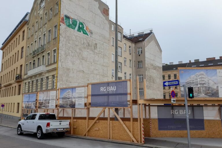leerer Bauplatz, davor Plakate und eine Absperrung aus Holz