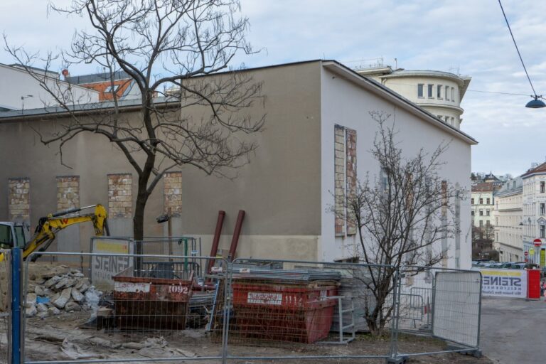 Gebäude mit vermauerten Fenstern, davor Bauzaun, Mulden und Bagger