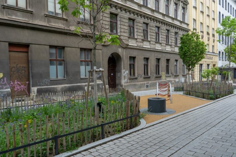 Grünfläche, Brunnen und Spielgerät in einer Fußgängerzone vor einer Häuserzeile